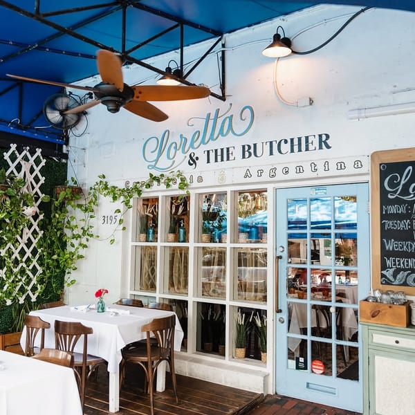 Outdoor dining area of Loretta & The Butcher, an Argentinian restaurant, with tables, plants, and a blue awning.