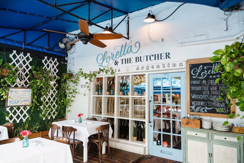 Outdoor dining area of Loretta & The Butcher, an Argentinian restaurant, with tables, plants, and a blue awning.