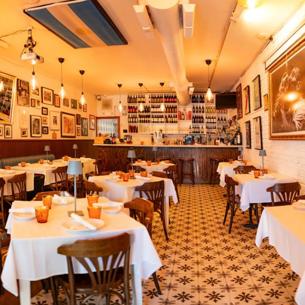 Cozy restaurant interior with white tablecloths, wooden chairs, a bar, and framed art on the walls.