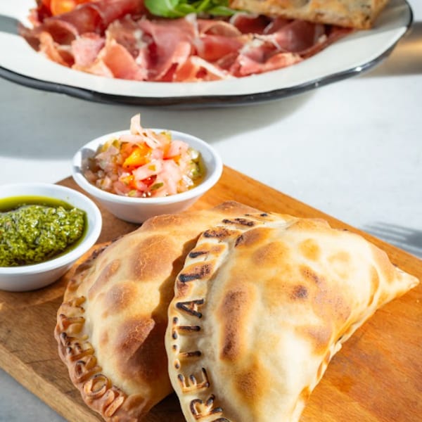 Two empanadas with dipping sauces on a wooden board, and a burrata salad in the background.
