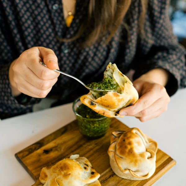 Person spoons green chimichurri sauce into a golden empanada on a wooden board.