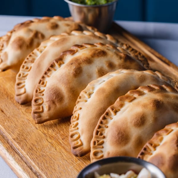Row of golden-brown empanadas with "LORENA" embossed on them, served on a wooden board with sauces.