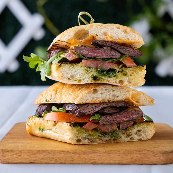 Two halves of a steak sandwich with pesto, tomatoes, and arugula, stacked on a wooden board.