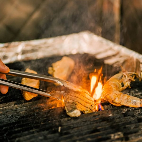 A hand uses tongs to flip shrimp and fish fillets cooking on a fiery grill.